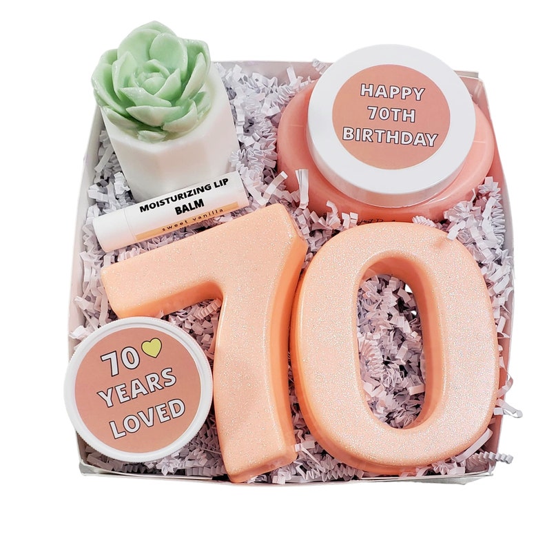 May include: A gift box filled with pink and white items for a 70th birthday celebration. The box includes a pink bath bomb with the text "Happy 70th Birthday", a pink lip balm, a small pink jar with the text "70 Years Loved", and two large pink numbers "70".