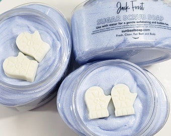 Op de afbeelding: Drie doorzichtige potten gevuld met lichtblauwe suikerscrubzeep. Elke pot bevat witte zeepstukjes in de vorm van wanten. De potten zijn gelabeld met "Jack Frost Sugar Scrub Soap" en "sunbasilsoap.com".
