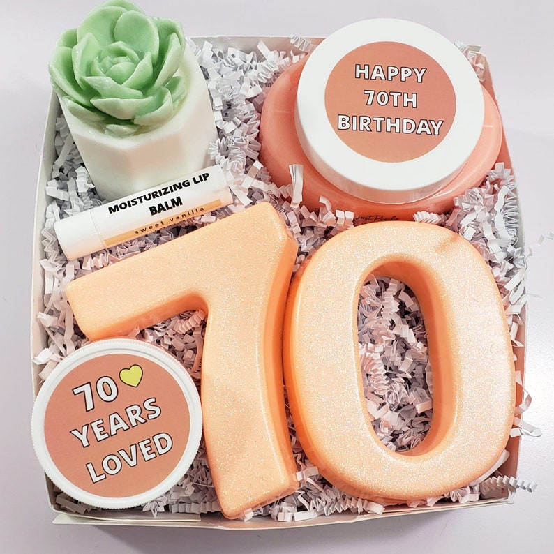May include: A gift box filled with items for a 70th birthday celebration. The box contains a pink bath bomb, a lip balm, a small succulent plant, and two large orange bath bombs shaped like the numbers 7 and 0. The bath bombs are decorated with glitter. The box is filled with white shredded paper. The lid of the box has a pink circle with the text "Happy 70th Birthday".