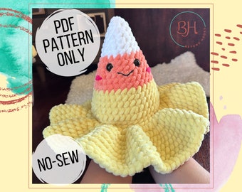 May include: A crochet pattern for a no-sew candy corn plush toy. The pattern is available as a PDF download. The image shows a completed plush toy with a yellow, orange, and white color scheme. The toy has a smiling face with black eyes and a pink mouth.