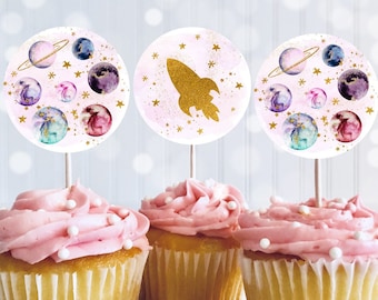 May include: Three pink frosted cupcakes with white sprinkles topped with round paper cupcake toppers featuring a watercolor galaxy theme. One topper has a gold rocket, another has a gold star, and the third has a gold planet with a ring.