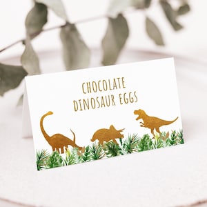 May include: White card with gold text that reads "Chocolate Dinosaur Eggs". The card features a gold silhouette of three dinosaurs, including a T-Rex, a Triceratops, and a Brachiosaurus, surrounded by green foliage.
