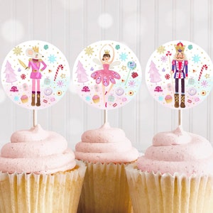May include: Three pink frosted cupcakes with round, white cupcake toppers featuring illustrations of a ballerina, a nutcracker, and a mouse. The toppers are decorated with snowflakes, candy canes, and other festive details.