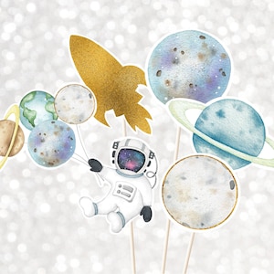 May include: A set of astronaut-themed party decorations featuring a white astronaut with a galaxy print on the helmet, a gold rocket, and five planets with rings. The planets are painted in shades of blue, green, and brown.
