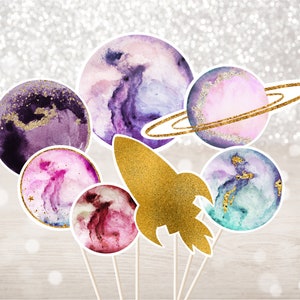 May include: A set of seven watercolor painted planets and a rocket ship on sticks. The planets are painted in shades of purple, pink, blue, and green with gold glitter accents. The rocket ship is gold with glitter accents.