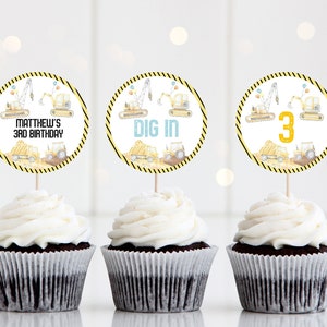 May include: Three cupcakes decorated with round construction vehicle themed cupcake toppers. The toppers feature yellow construction vehicles, a blue sky, and yellow and black striped borders. The toppers say "Matthew's 3rd Birthday", "Dig In", and "3".