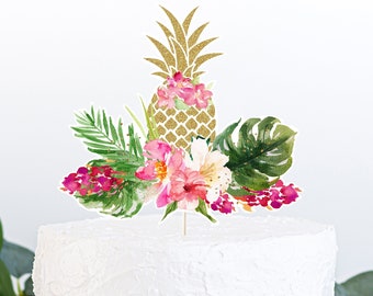 May include: A white frosted cake topped with a gold glitter pineapple and tropical flowers. The cake topper includes pink, red, and white flowers, and green leaves. The cake sits on a wooden cake stand.