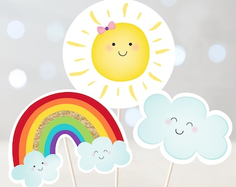 Puede incluir: Tres adornos para pasteles coloridos que representan un sol amarillo con un lazo rosa, un arcoíris con purpurina dorada y una nube azul con una cara sonriente.