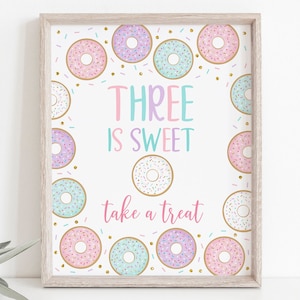 Op de afbeelding: Een ingelijste print met een witte achtergrond, met kleurrijke donuts en de tekst "THREE IS SWEET take a treat". De donuts zijn roze, blauw, paars en wit met hagelslag en gouden accenten. De lijst is van licht hout.