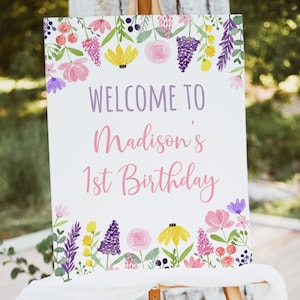 May include: A white sign with a floral border and the text "Welcome to Madison's 1st Birthday" in purple and pink lettering.