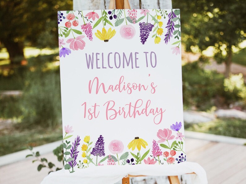 May include: A white sign with a floral border and the text "Welcome to Madison's 1st Birthday" in purple and pink lettering.