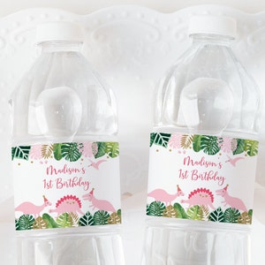 Pu&ograve; includere: Due bottiglie d'acqua in plastica trasparente con etichette personalizzate. Le etichette presentano un tema tropicale rosa e verde con dinosauri e il testo "Madison's 1st Birthday".