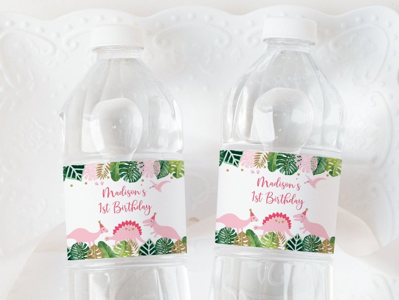 Pu&ograve; includere: Due bottiglie d'acqua in plastica trasparente con etichette personalizzate. Le etichette presentano un tema tropicale rosa e verde con dinosauri e il testo "Madison's 1st Birthday".