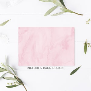 May include: A rectangular card with a soft pink watercolor design. The text "INCLUDES BACK DESIGN" is printed at the bottom. The card is surrounded by white flowers, green leaves, and a wooden clothespin on a white wooden surface.