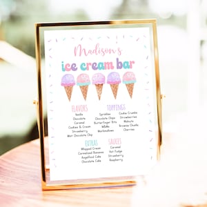 May include: A framed sign with the text "Madison's ice cream bar" in pink and blue. The sign lists ice cream flavours, toppings, extras, and sauces, with illustrations of ice cream cones. The frame is gold.
