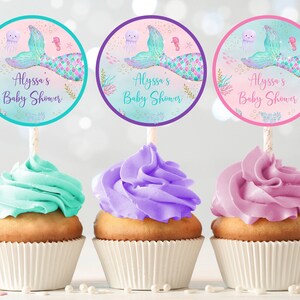 May include: Three cupcakes decorated with turquoise, purple, and pink frosting. Each cupcake has a round, colorful sticker with a mermaid tail, a jellyfish, and the text "Alyssa's Baby Shower".