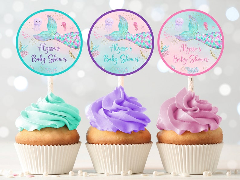 May include: Three cupcakes decorated with turquoise, purple, and pink frosting. Each cupcake has a round, colorful sticker with a mermaid tail, a jellyfish, and the text "Alyssa's Baby Shower".
