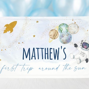 May include: A white and blue space-themed baby shower sign with gold glitter stars. The sign features an astronaut holding planets as balloons, a rocket ship, and the text "MATTHEW'S first trip around the sun".