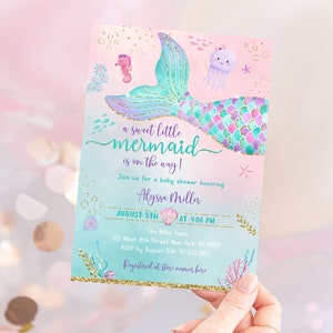 May include: A baby shower invitation with a mermaid tail design. The invitation is pink and blue with gold glitter accents. The text reads "a sweet little mermaid is on the way!" and includes the date, time, and location of the shower.