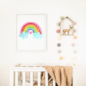 May include: A white crib with a brown blanket and a framed watercolor painting of a rainbow with clouds and a pink heart above it. A string of colorful light bulbs hangs on the wall above the crib.
