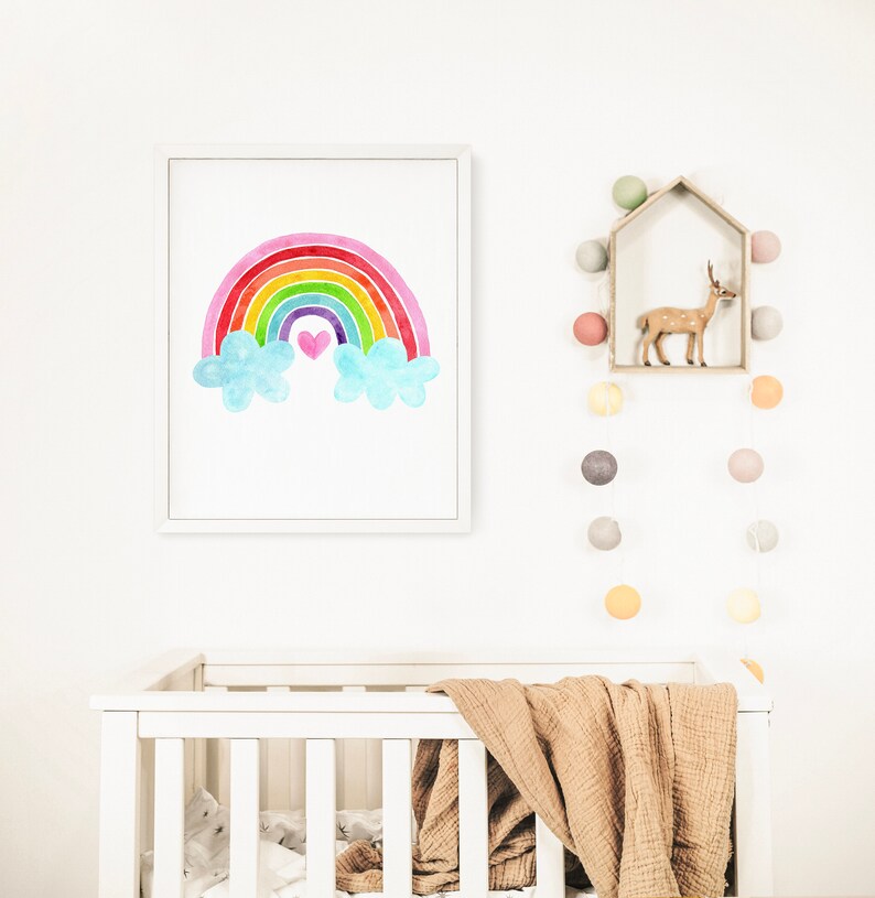 May include: A white crib with a brown blanket and a framed watercolor painting of a rainbow with clouds and a pink heart above it. A string of colorful light bulbs hangs on the wall above the crib.