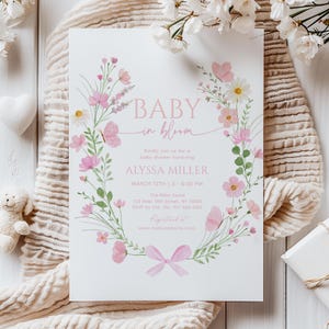 May include: A baby shower invitation with a floral wreath design. The invitation features the text "BABY in bloom" in pink, along with event details. Pink flowers and butterflies decorate the invitation, which is on a white background.