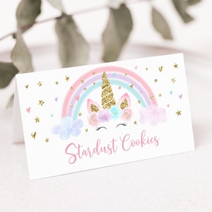 May include: A white card with a unicorn design and the text "Stardust Cookies." The unicorn has a gold glitter horn and is set against a rainbow with pastel colors. Gold glitter stars and hearts are scattered around the design.