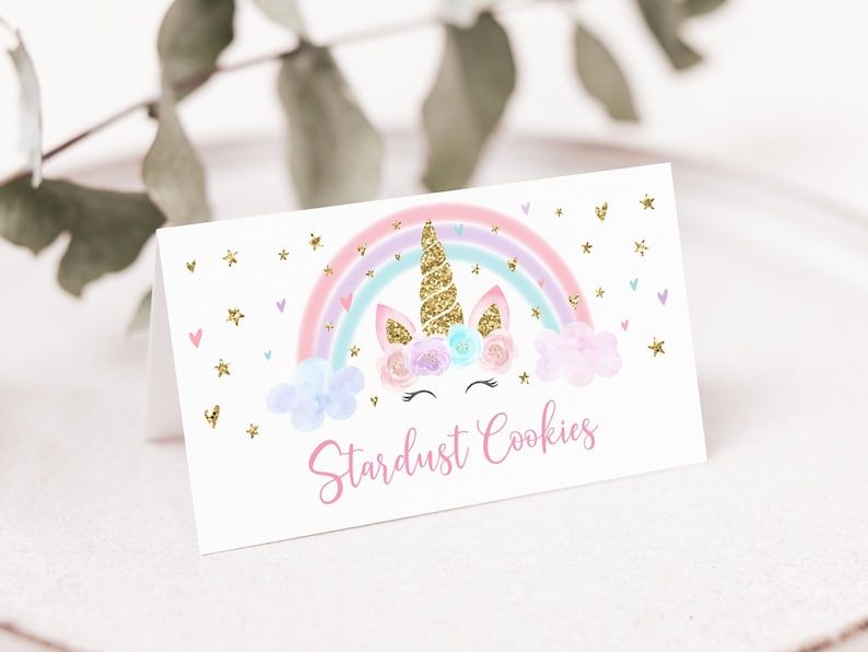 May include: A white card with a unicorn design and the text "Stardust Cookies." The unicorn has a gold glitter horn and is set against a rainbow with pastel colors. Gold glitter stars and hearts are scattered around the design.