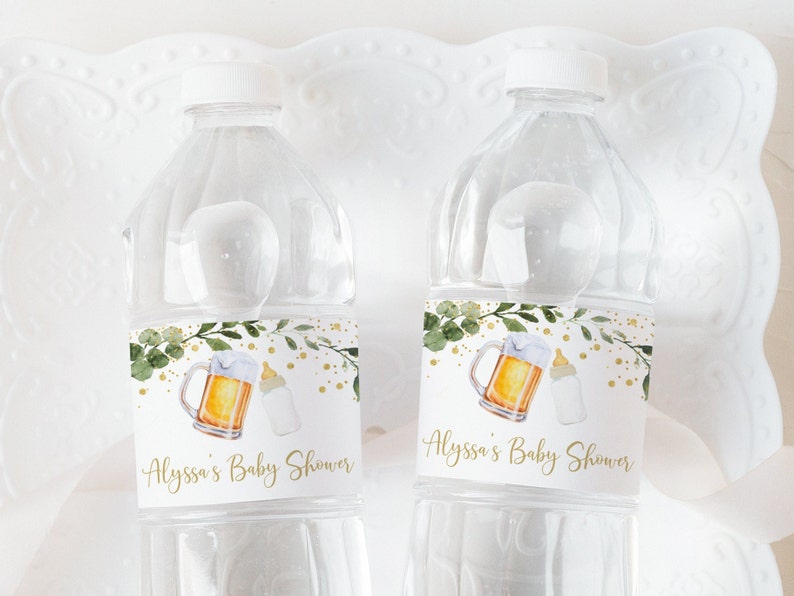 May include: Two clear plastic water bottles with white caps. Each bottle has a label with a watercolor illustration of a beer mug and a baby bottle, with the text "Alyssa's Baby Shower" in gold script. Green leaves and gold dots decorate the label.