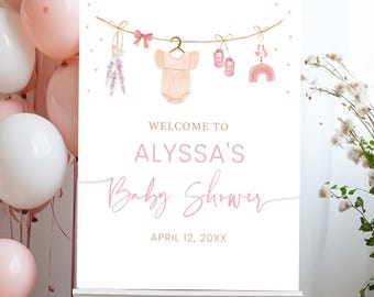 Op de afbeelding: Een wit welkomstschild voor een babyshower met roze en gouden accenten. Het bord heeft een string met babyartikelen, waaronder een rompertje, slofjes en een regenboog. De tekst op het bord luidt "Welkom op Alyssa's babyshower 12 april 20XX".