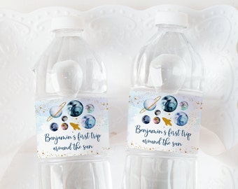 May include: Two clear plastic water bottles with blue and gold labels. The labels feature a watercolor design of planets and stars with the text "Benjamin's first trip around the sun".