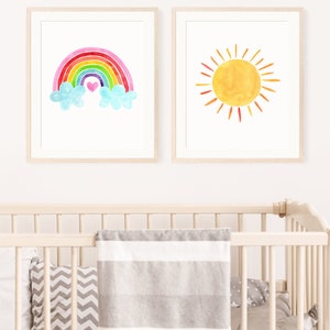 May include: Two framed watercolor prints, one with a rainbow and clouds and the other with a yellow sun. The rainbow is multi-colored with pink, red, orange, yellow, green, blue, and purple. The sun has orange rays.