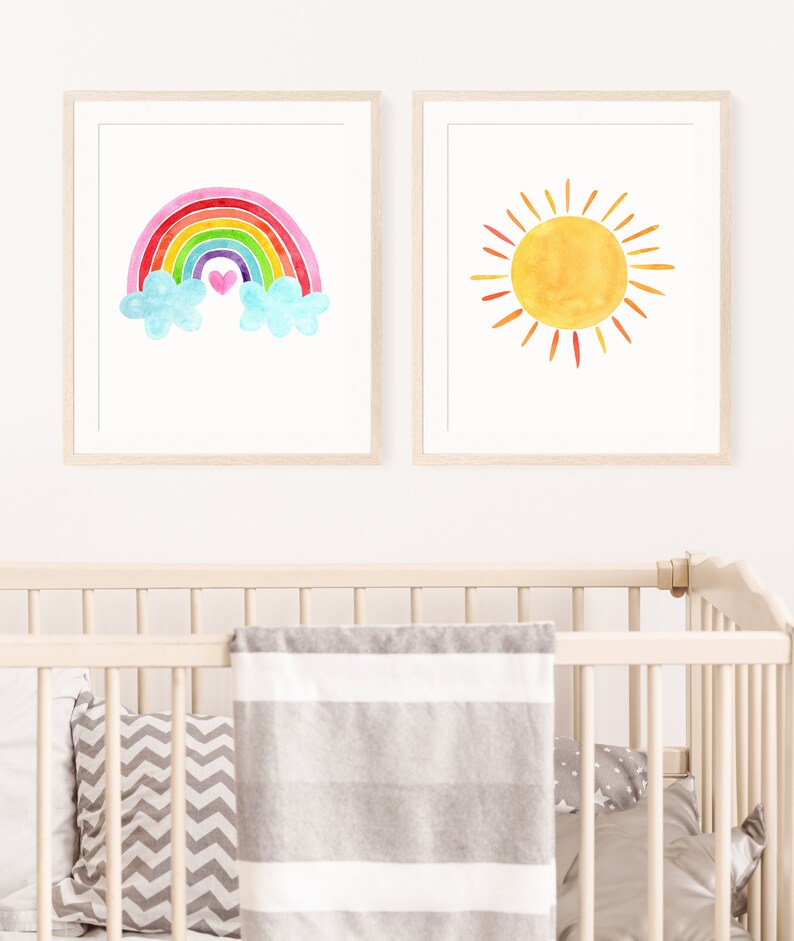 May include: Two framed watercolor prints, one with a rainbow and clouds and the other with a yellow sun. The rainbow is multi-colored with pink, red, orange, yellow, green, blue, and purple. The sun has orange rays.