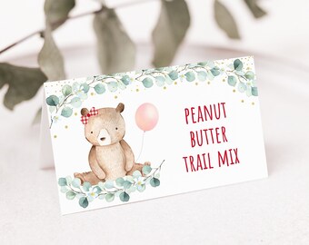 May include: A white card with a brown bear holding a pink balloon. The bear is wearing a red and white bow. The card has green leaves and gold glitter around the edges. The text "PEANUT BUTTER TRAIL MIX" is printed in red on the card.