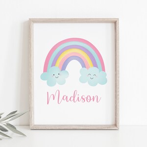 Puede incluir: Un arco&iacute;ris pastel con dos nubes sonrientes y el nombre Madison escrito en rosa debajo. El arco&iacute;ris est&aacute; sobre un fondo blanco con un marco de madera claro.