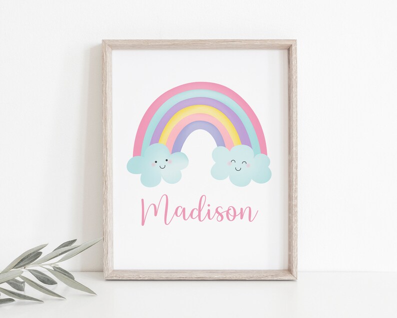 Puede incluir: Un arco&iacute;ris pastel con dos nubes sonrientes y el nombre Madison escrito en rosa debajo. El arco&iacute;ris est&aacute; sobre un fondo blanco con un marco de madera claro.