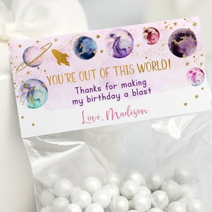 Puede incluir: Una bolsa de regalo rosa y morada con un diseño de temática espacial. La bolsa tiene una etiqueta que dice "You're out of this world! Thanks for making my birthday a blast. Love, Madison."