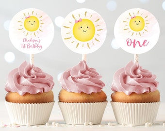 May include: Three pink frosted cupcakes with white cupcake liners. Each cupcake has a round, white paper topper with a yellow sun with a pink bow and gold glitter. The toppers say "Madison's 1st Birthday", "one", and a sun with a pink bow.