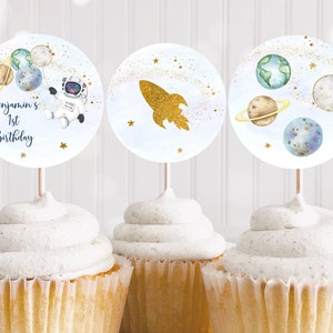 Op de afbeelding: Drie cupcakes met ronde toppers met een ruimtethema. Eén topper heeft een astronaut en planeten, een andere een gouden raket en de laatste planeten. De eerste topper leest "Benjamin's 1st Birthday".