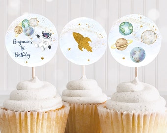 May include: Three cupcakes topped with round toppers featuring a space theme. One topper has an astronaut and planets, another a gold rocket, and the last has planets. The first topper reads "Benjamin's 1st Birthday".