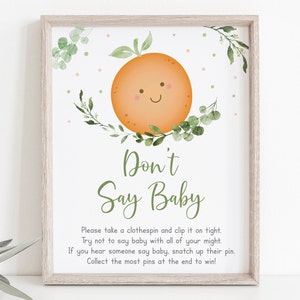 Puede incluir: Un cartel imprimible para un juego de baby shower. Presenta una naranja de dibujos animados con una cara sonriente y hojas verdes. El texto dice "No digas bebé" e incluye instrucciones para el juego.