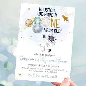 May include: A blue and gold astronaut-themed first birthday party invitation with a cartoon astronaut holding balloons shaped like planets. The text reads "Houston, we have a one year old!" and "Join us to celebrate Benjamin's 1st trip around the sun".