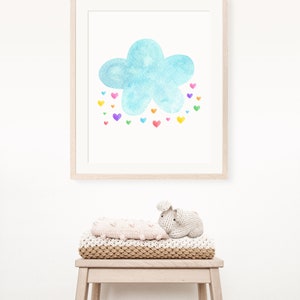 May include: A framed watercolor painting of a blue cloud with a rainbow of hearts falling from it. The painting is hanging on a white wall above a wooden stool with a knitted blanket and a crocheted bunny toy.