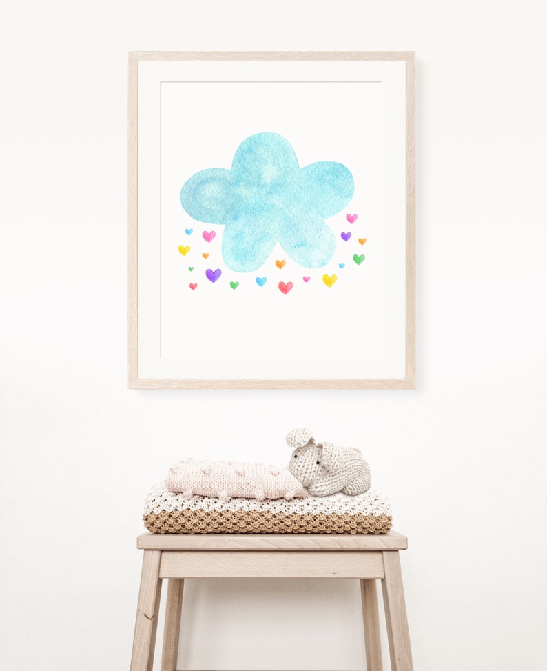May include: A framed watercolor painting of a blue cloud with a rainbow of hearts falling from it. The painting is hanging on a white wall above a wooden stool with a knitted blanket and a crocheted bunny toy.