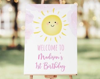 May include: A white welcome sign with a pink watercolor background and a yellow sun with a smiling face. The text on the sign reads "Welcome to Madison's 1st Birthday".
