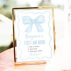 Puede incluir: Un cartel blanco con un lazo azul de acuarela en la parte superior. El cartel dice "Benjamin's First ABC Book" y tiene instrucciones para elegir una letra, decorar y firmar tu nombre. Termina con "¡Gracias!"