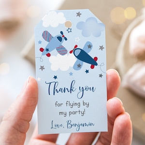 May include: Blue and red airplane thank you tag with silver glitter stars and clouds. The tag says "Thank you for flying by my party! Love, Benjamin".