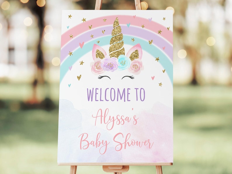 May include: A welcome sign for a baby shower with a unicorn illustration. The unicorn has a gold horn and a floral crown. The sign has a rainbow and clouds with the text "Welcome to Alyssa's Baby Shower".