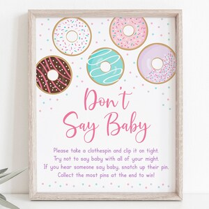 May include: A pink and white sign with the text "Don't Say Baby" and a graphic of five donuts with pink, blue, and brown frosting. The sign is framed in a light wood frame.