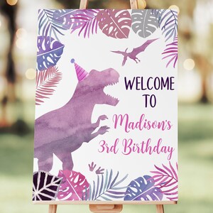 May include: A welcome sign for a 3rd birthday party with a purple dinosaur wearing a pink party hat. The sign has a watercolor background with pink, purple, and blue tropical leaves. The text on the sign reads "Welcome to Madison's 3rd Birthday".